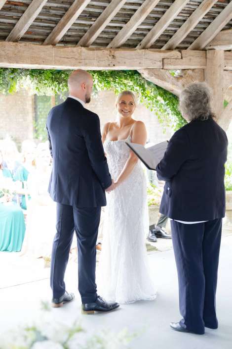 bride and groom exchange vows A bride in a white lace gown and a groom in a navy suit hold hands as an officiant reads from a book during their outdoor wedding ceremony under a wooden canopy.