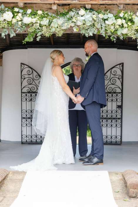 outdoor wedding ceremony under floral arch Bride and groom stand facing each other, hands joined, as an officiant presides under a lush floral arch.