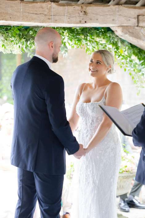 bride and groom exchange vows Bride in a lace white dress and groom in a navy suit hold hands and smile during an outdoor wedding ceremony.