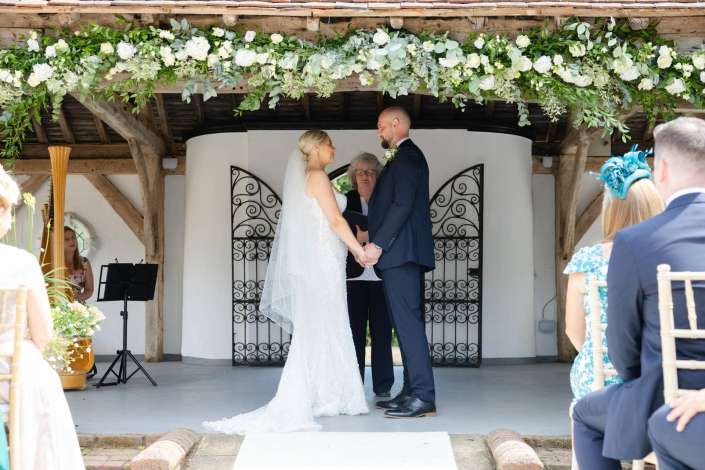 wedding ceremony under floral arch Bride and groom exchange vows outdoors beneath a lush white-and-green floral arch with an officiant standing between them.