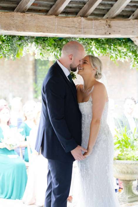 intimate wedding moment Bride and groom kiss while holding hands under a rustic, greenery-adorned wooden beam at their outdoor wedding.