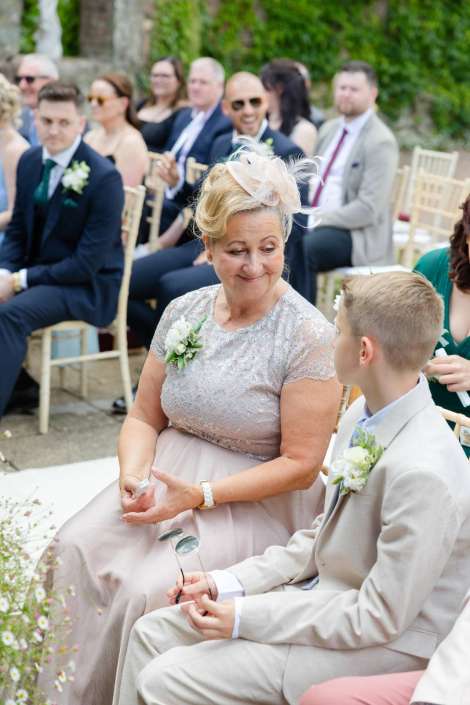mother and son at wedding An older woman in a lavender lace dress with a fascinator sits next to a young man in a light gray suit at an outdoor wedding.