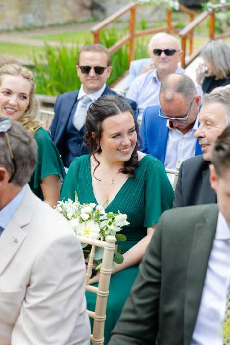 emerald green bridesmaid with bouquet A smiling woman in a green dress sits among wedding guests outdoors, holding a white bouquet.