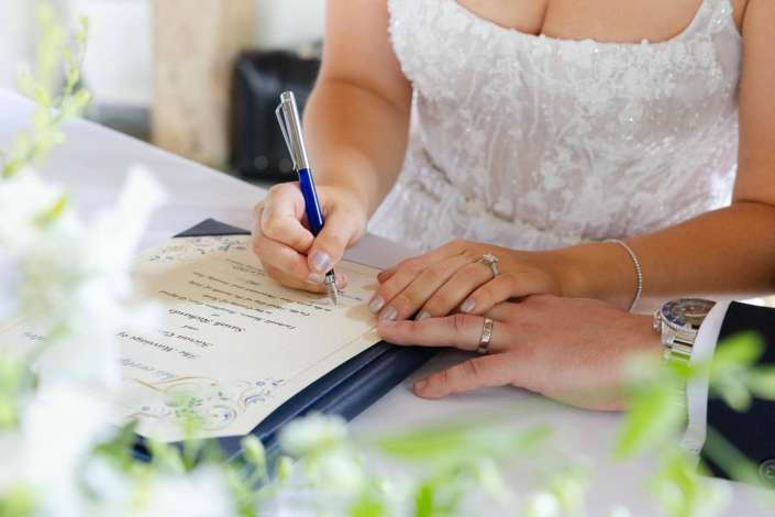 bride signing marriage certificate A bride signs a document with a blue pen, her lace wedding dress visible, with wedding rings on her fingers and a groom’s hand nearby.