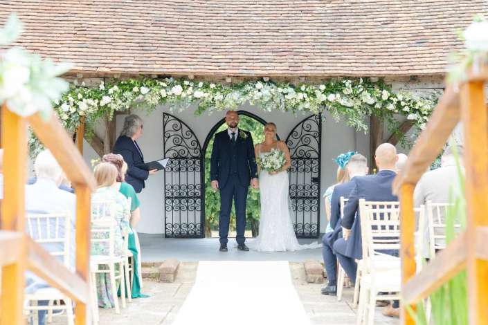 outdoor wedding ceremony under floral arch Bride and groom stand hand in hand under a flower-covered arch at an outdoor wedding ceremony, with guests seated along a white aisle.