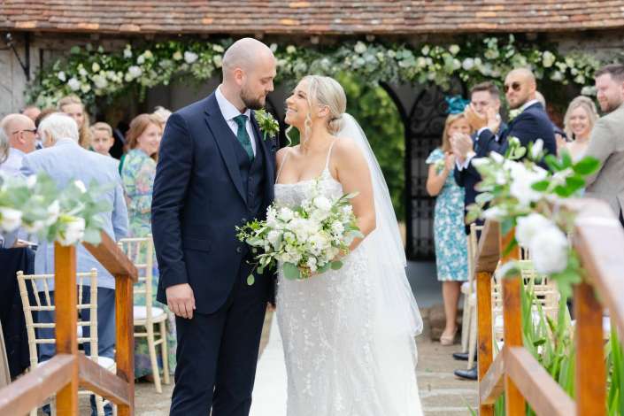newlyweds walking down aisle Bride and groom smile at each other in an outdoor floral aisle as guests applaud in the background.