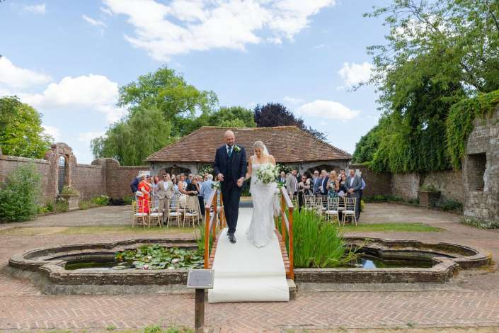outdoor wedding aisle walk Bride and groom walk down a white aisle runner at an outdoor garden wedding, surrounded by seated guests and a lily-filled pond.