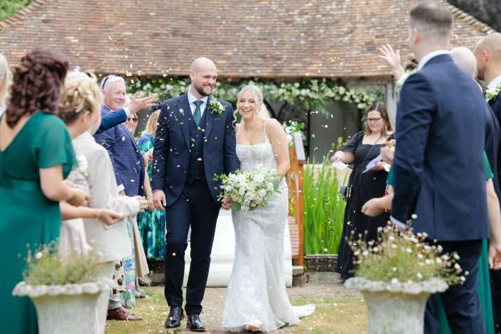 newlyweds walk through confetti A bride in a white wedding dress and a groom in a navy suit walk down a garden aisle while guests shower them with gold confetti.