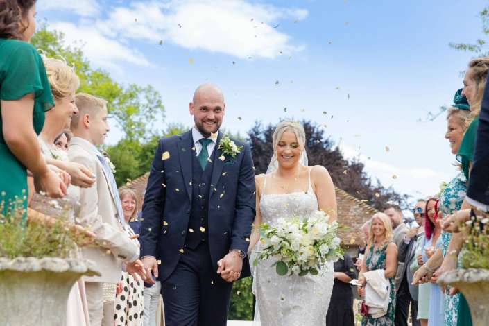 bride and groom walk amid confetti A smiling bride in a white wedding gown and a groom in a navy suit walk hand in hand down an outdoor aisle as guests on both sides shower them with gold confetti.