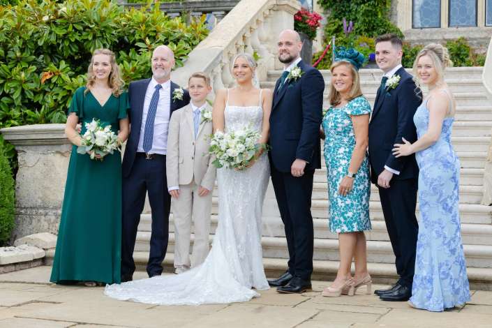 wedding party on stone steps Bride in a white lace gown and groom in a navy suit pose with their wedding party on stone steps, surrounded by family in green and blue dresses and suits.