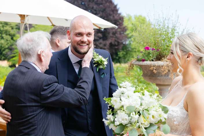 groom embraced by father An outdoor wedding moment with a smiling groom in a navy suit as an older man tenderly touches his face, while the bride in a white dress holds a bouquet nearby.