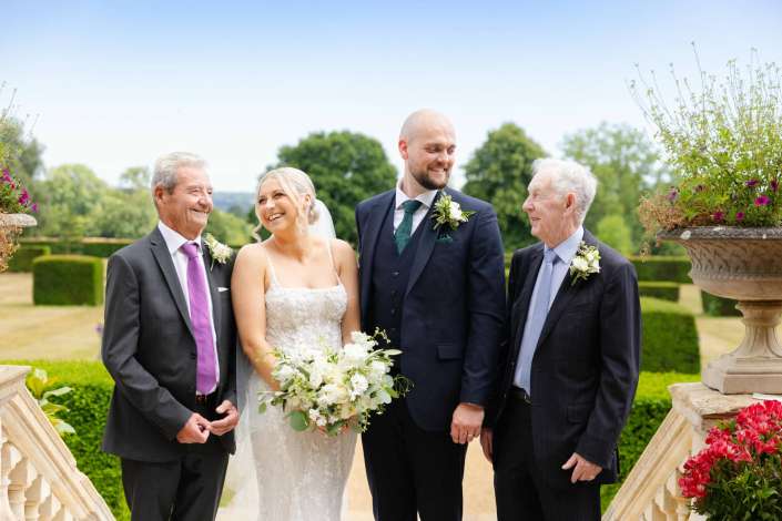 bride and groom with fathers Smiling bride in a white lace wedding dress holds a bouquet as she stands with her groom and two older men in suits in a sunny formal garden.