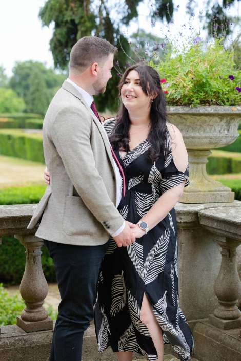 romantic garden couple portrait A couple stands hand in hand, smiling at each other beside a stone balustrade with a large flower-filled planter in a garden.