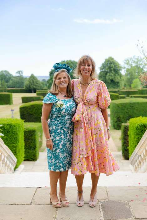 two women in floral dresses Two smiling women in pastel floral dresses stand together on a stone staircase in a formal garden with neatly trimmed hedges.