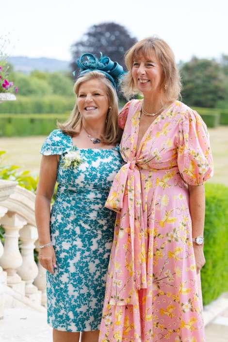 two friends in floral dresses Two women in floral dresses pose outdoors, smiling, with one in a teal dress and blue fascinator and the other in a pink dress with a waist tie.