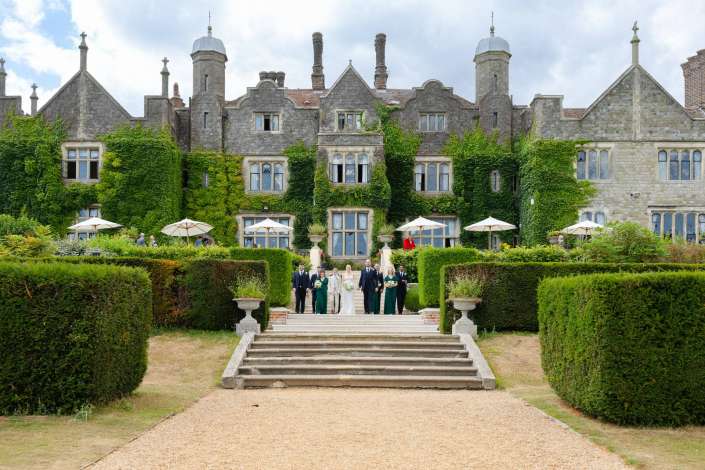 ivy covered manor wedding party A wedding party poses on the stone steps in front of an ivy-covered historic manor with a formal garden.