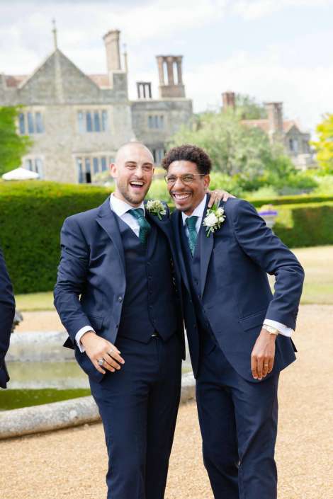 two grooms in navy suits Two men in navy suits with green ties and boutonnieres stand together, smiling with arms around each other in a formal garden.