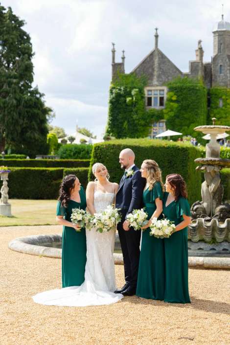 elegant garden wedding party Bride in a white wedding gown and groom in a dark suit pose with three bridesmaids in emerald green dresses, all holding white bouquets, in a sunlit formal garden beside a stone fountain.