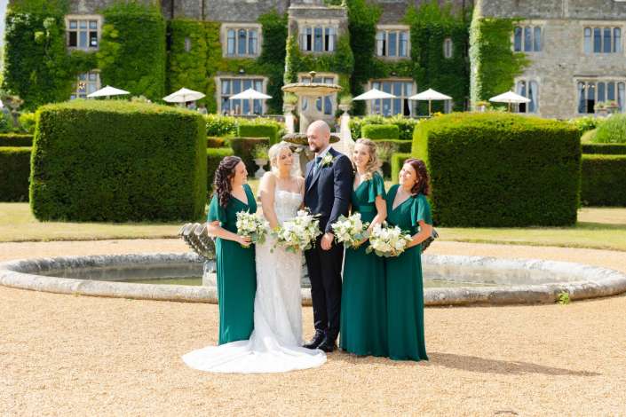 garden bridal party portrait Bride and groom stand with four bridesmaids in emerald dresses holding white bouquets in front of a fountain in a formal garden.