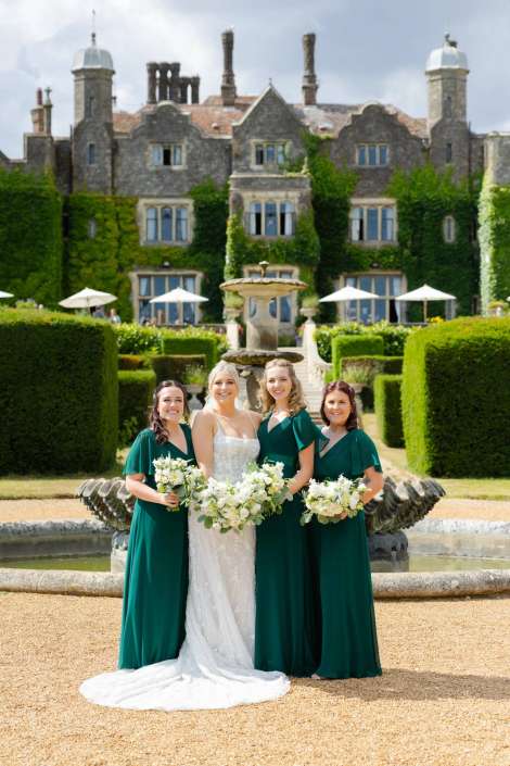 elegant garden wedding party Bridal party standing in a formal garden in front of an ivy-covered mansion and fountain; the bride in a white gown flanked by three bridesmaids in emerald green dresses holding white bouquets.