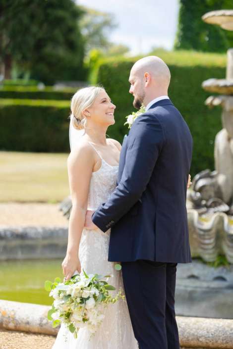 bride and groom embrace Bride in a white wedding dress and veil holds a bouquet and smiles at her groom in a navy suit outdoors beside a fountain.