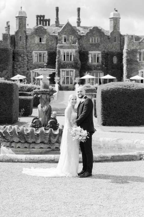 bride and groom at manor Bride and groom in wedding attire pose in front of a grand ivy-covered mansion with a fountain and formal gardens.