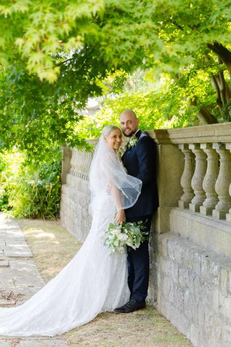 bride and groom by balustrade Bride in a white wedding dress and veil stands with a groom in a dark suit beside a stone balustrade under leafy trees.