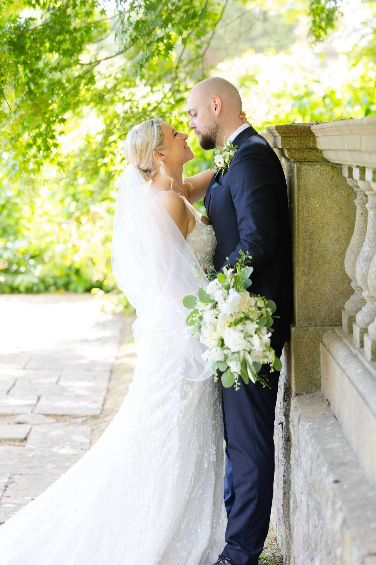 bride and groom embrace Bride in a white gown and veil and a groom in a navy suit embrace beside a stone railing in a sunlit garden, with the bride holding a white bouquet.