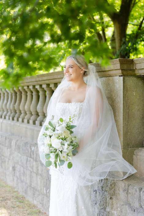 bride with white bouquet Smiling bride in a white wedding gown with veil, holding a white bouquet outdoors beside a stone balustrade.