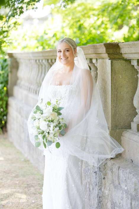 bride in white lace veil Smiling bride in a white wedding gown with veil stands beside a stone balustrade in a sunlit garden, holding a white bouquet with greenery.