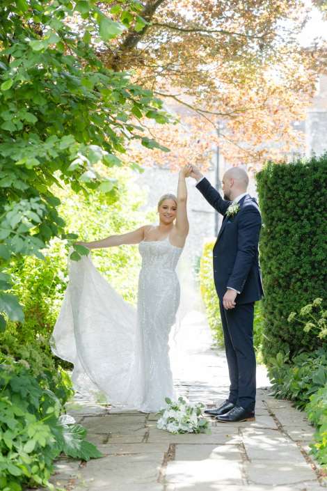bride and groom dancing outdoors A bride in a white wedding dress and a groom in a dark suit dance on a sunlit garden path, with the bride holding up her train.