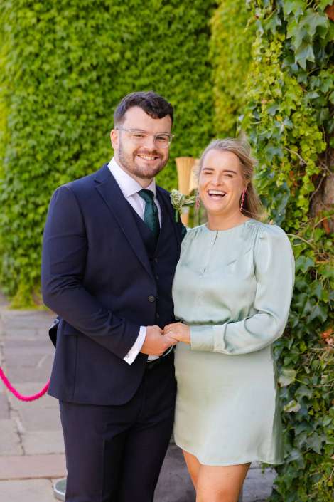 smiling couple in formal attire Smiling couple outdoors, the man in a navy suit with a green tie and boutonniere and the woman in a pale green dress, standing together against a lush green hedge.