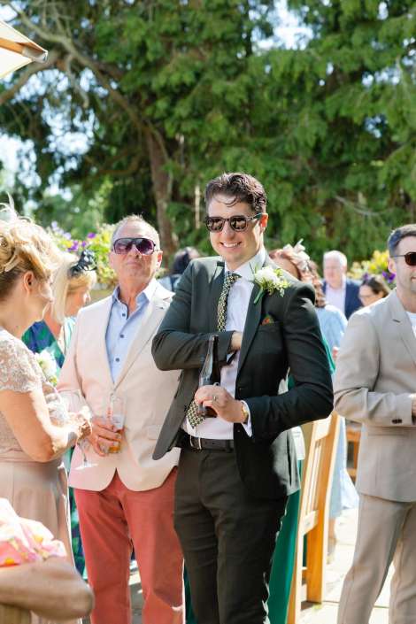 groom in green suit at wedding A man in a dark green suit and sunglasses smiles while holding a beer bottle at an outdoor wedding with guests.