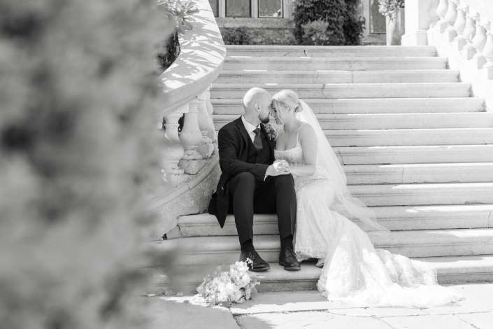 wedding portrait on marble steps Bride in a wedding gown and veil and groom in a suit sit close on a stone staircase, leaning their heads together and holding hands.