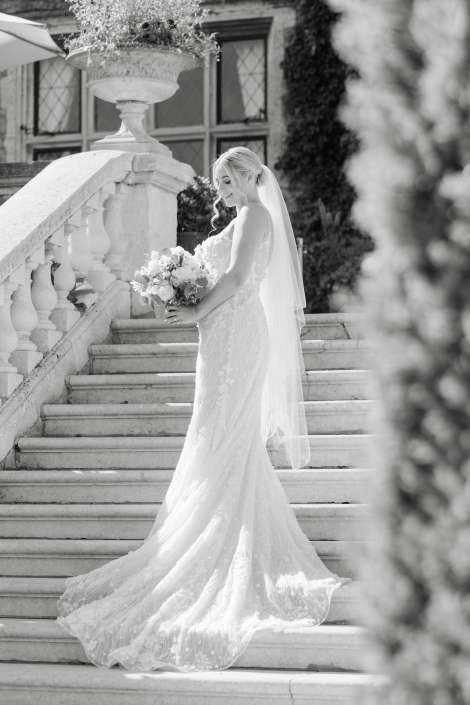 bride on marble staircase Bride in a lace wedding gown with a long train and veil stands on a stone staircase, holding a bouquet.