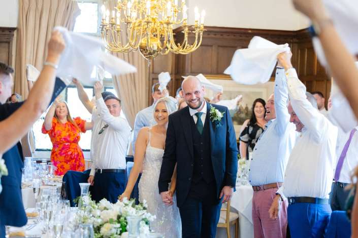 joyful wedding entrance Bride and groom walk hand in hand through a room full of guests waving napkins at their wedding reception.