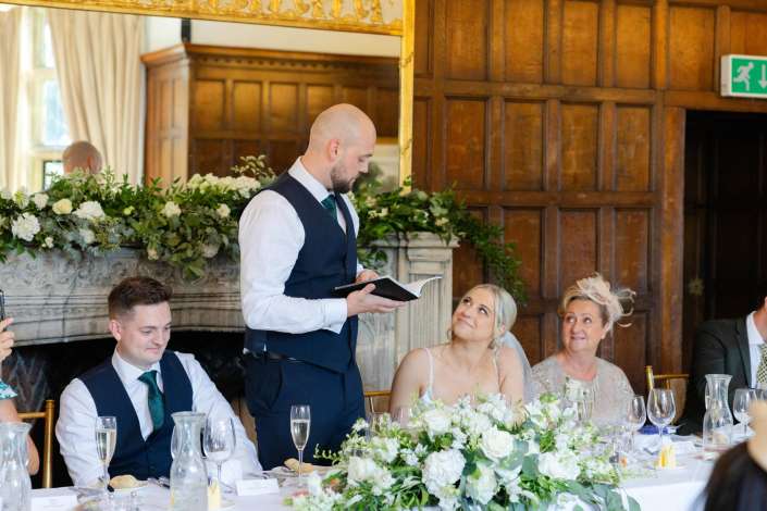 wedding reception speech A bald man in a navy vest delivers a speech at a wedding reception table decorated with white flowers as guests listen.