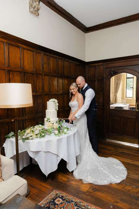 bride and groom cutting cake Bride and groom cut a white tiered wedding cake on a table decorated with greenery in a wood-paneled room.