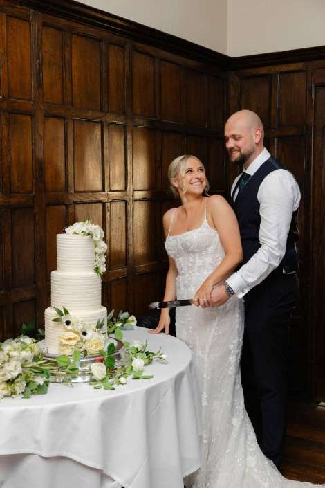 bride and groom cake cutting Bride in a lace wedding dress and groom in a vest cut a tall white wedding cake decorated with flowers on a table beside dark wooden paneling.