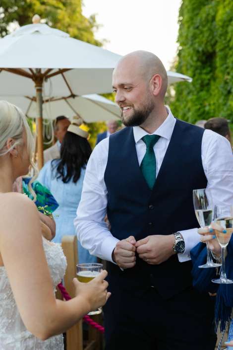 garden wedding guests chatting Bearded man in a navy vest and green tie talks with a bride in a white dress at an outdoor wedding, both holding drinks.