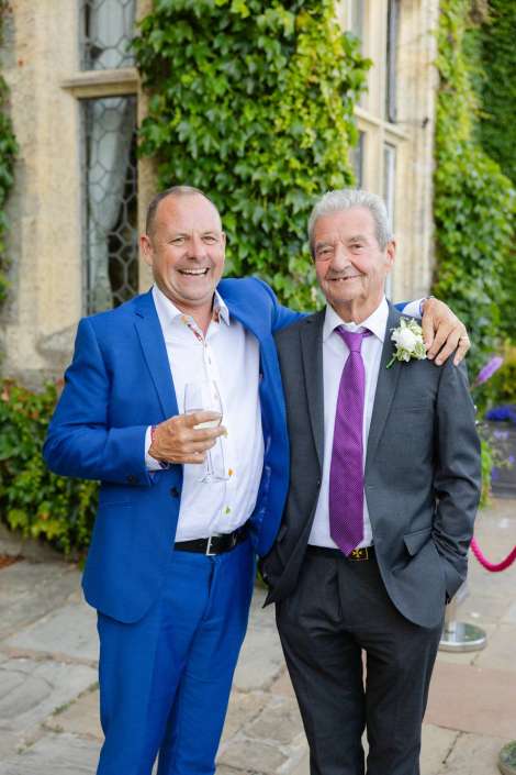 friends in suits at wedding Two men in suits stand together smiling outdoors, one in a bright blue suit holding a wine glass.