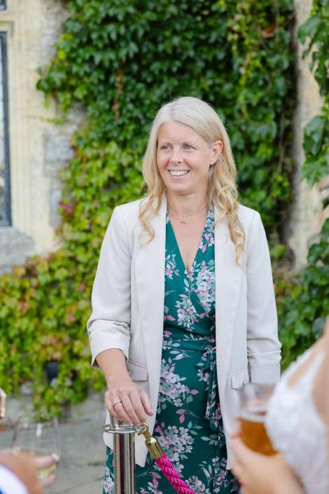 smiling woman in floral dress Smiling blonde woman in a cream blazer and teal floral dress stands outdoors with an ivy-covered wall behind her.