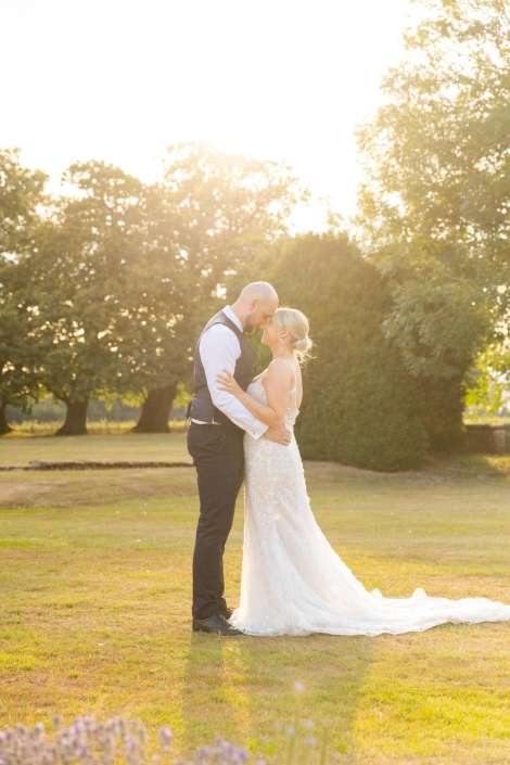 golden hour wedding embrace A bride in a white wedding gown and a groom in a dark vest embrace and kiss in a sunlit park.