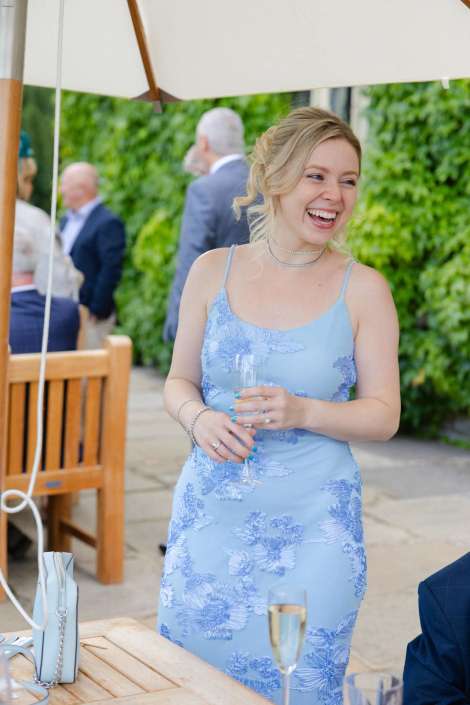 smiling woman in blue floral dress A smiling woman in a light blue floral dress holding a champagne flute at an outdoor garden party.