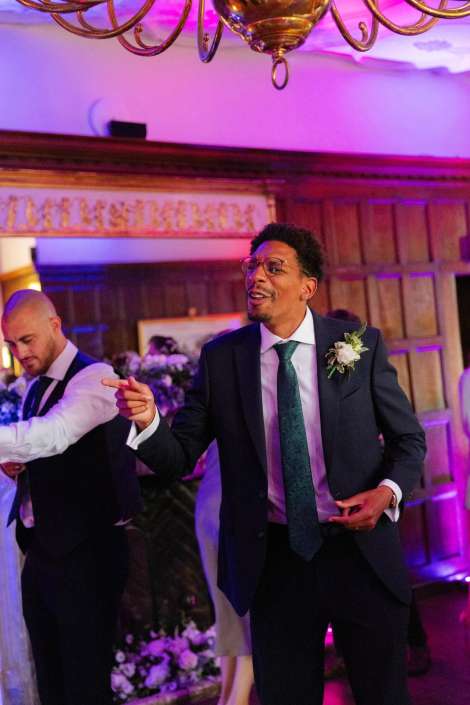 man in suit dancing at wedding A man in a dark suit with a boutonniere and green tie gestures while speaking at a wedding reception, with another guest in a vest in the background.