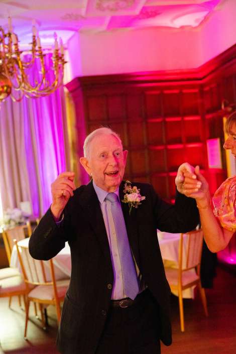joyful dancing at wedding reception An elderly man in a dark suit and light blue tie dances with a partner at a festive indoor event under pink and purple lighting.