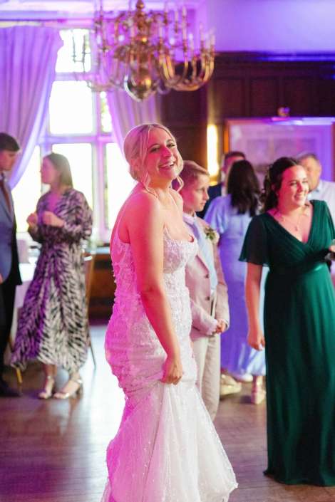 bride on the dance floor A smiling bride in a white lace wedding gown stands on a dance floor with guests at a reception under a chandelier.