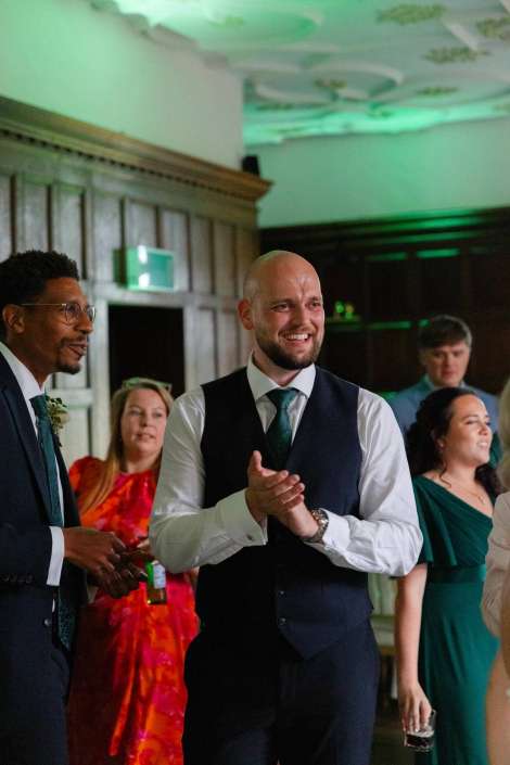 clapping guests at reception A smiling man in a white shirt and navy vest claps at a formal event, surrounded by other well-dressed guests.
