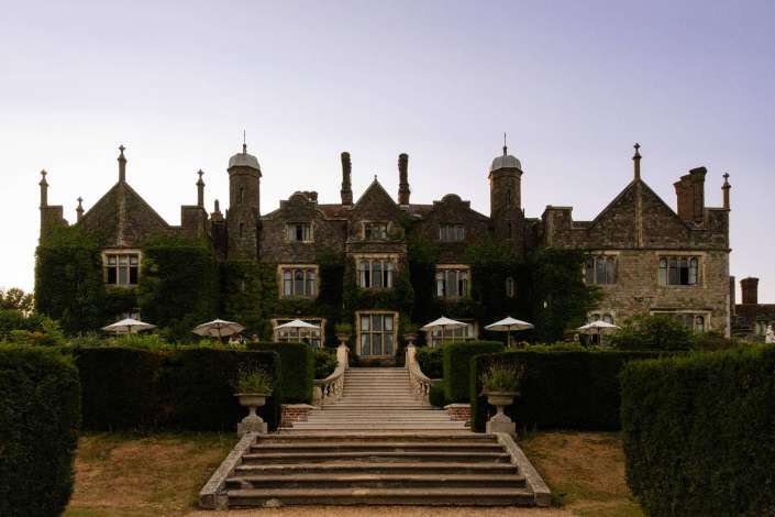 ivied manor house Ivied stone manor with a grand central staircase, numerous chimneys, and formal hedged gardens under a pale sky.