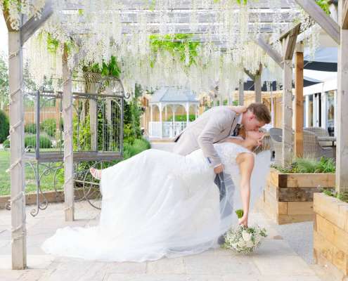 Bride and groom share a kiss as the groom dips the bride beneath a flowering wooden pergola. Photo by Louisa Dettmer Wedding Photography, Kent Wedding Photographer